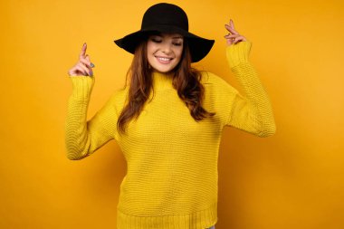 Sarı arka planda duran esmer bir kadın sarı bir kazak ve siyah şapkayla dans ediyor ve gülümsüyor..