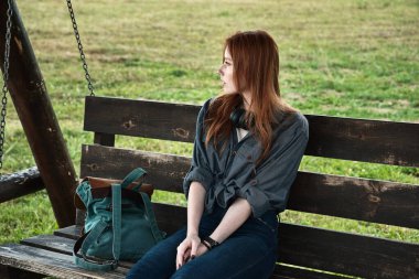 Kızıl saçlı bir kız, tahta bir bankta sırt çantasıyla oturur sallanır ve başını çevirir..