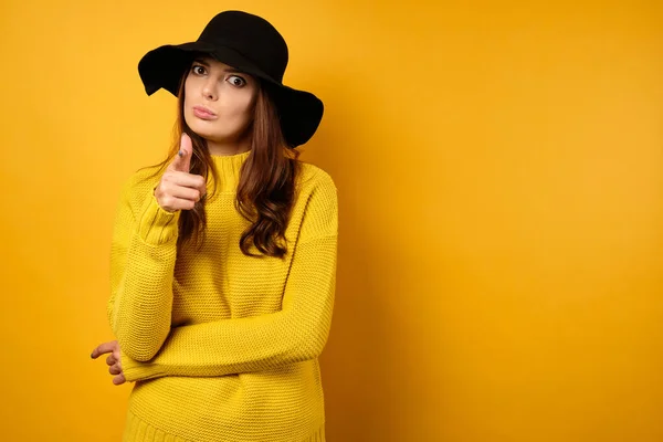 En vacker brunett i gul tröja och svart hatt står på en gul bakgrund rynkar pannan och pekar fingret på ramen — Stockfoto