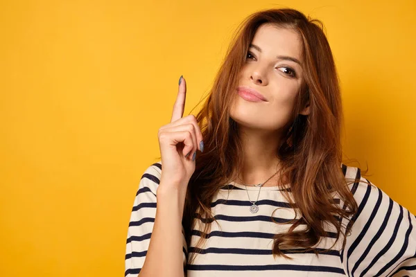 Eine schöne Brünette in einem gestreiften T-Shirt steht auf gelbem Hintergrund und hält ihren Zeigefinger in die Höhe. — Stockfoto