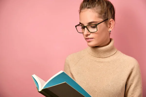 Una chica con cuello alto y gafas con el pelo recogido se levanta sobre un fondo rosa leyendo un libro . — Foto de Stock