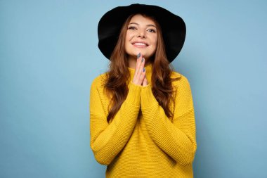 Esmer, mavi bir arka planda sarı bir süveter ve siyah bir şapkayla duruyor. Elleri önünde sıkıca bağlı, kameraya gülümsüyor.