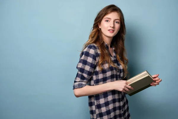 Светловолосая девушка в клетчатой рубашке стоит на синем фоне в полуобороте с открытой книгой и смотрит в рамку . — стоковое фото