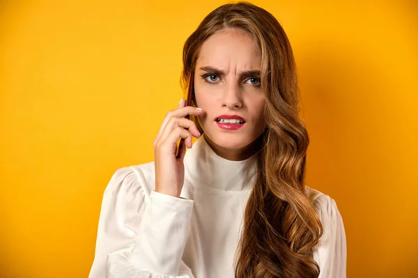 Uma menina bonita com lábios vermelhos fica em um fundo amarelo, falando ao telefone com uma careta descontente . — Fotografia de Stock