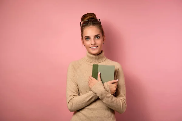 광선 이 높은 한 소녀는 스웨터 에 안경을 머리에 쓴 채 핑크 색 배경을 가지고 서서 책을 가슴에 쥐고 있다. — 스톡 사진
