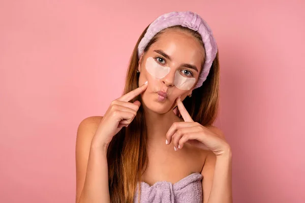 Portrait of a grimacing girl with a bandage on her head, in a lilac towel and with patches under her eyes on a pink background. — Stock Photo, Image