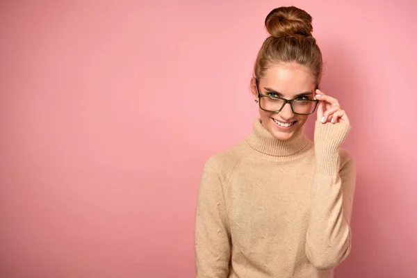 En flicka med en hög stråle står i en tröja på en rosa bakgrund, ler och tittar åt sidan, håller glasögon med handen — Stockfoto