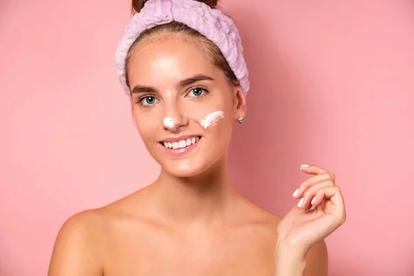 Seorang gadis dengan kulit bersih berdiri di atas latar belakang merah muda, tersenyum di depan kamera, dengan noda krim di wajahnya . — Stok Foto