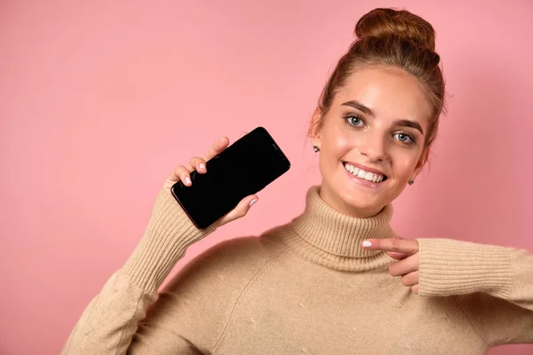 A girl with clean skin and a high bun joyfully points to the smartphone with her finger and looks at the frame. — Stock Photo, Image