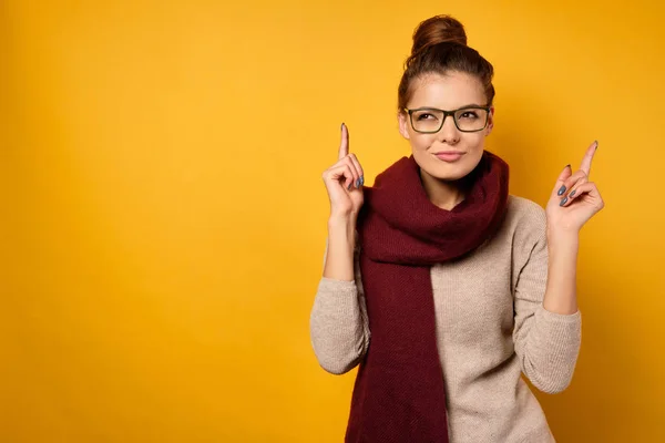 Ein Mädchen in Schal und Brille mit Fernlicht zeigt mit verschmitztem Gesichtsblinzeln auf gelbem Hintergrund die Finger nach oben. — Stockfoto