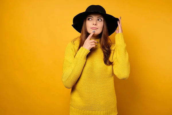 Brunetten i gul tröja och svart hatt ser tankfullt åt sidan med fingret mot läpparna. — Stockfoto
