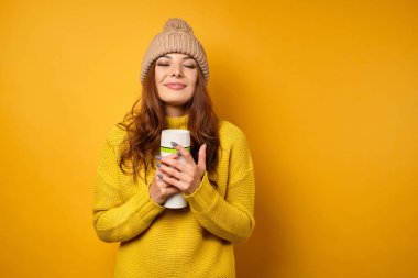 Sarı süveterli ve şapkalı bir esmer sarı arka planda duruyor, gözlerini kısarak, göğsüne termofiyi bastırıyor.