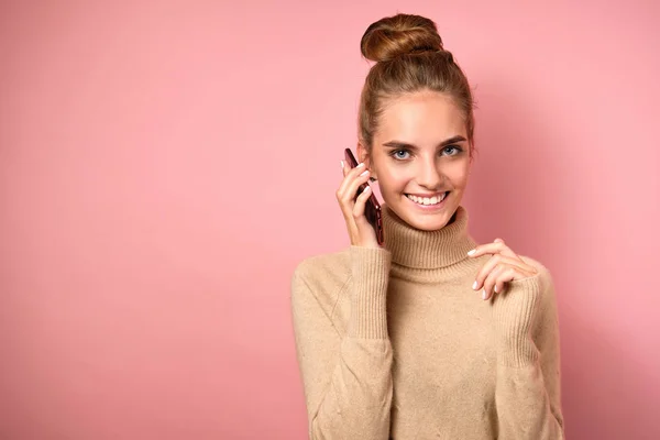터틀넥에서 높은 빔을 머리에 얹고 있는 소녀가 핸드폰을 귀에 가지고 와서는 핑크 색 배경에 미소를 짓습니다. — 스톡 사진