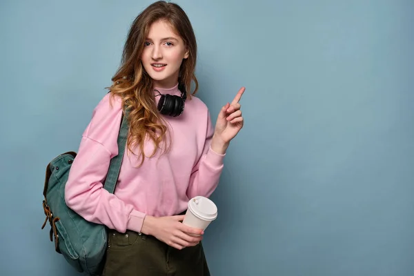 En flicka med ryggsäck, hörlurar på halsen står på en blå bakgrund med en papperskopp och pekar fingret åt sidan. — Stockfoto