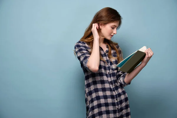 Egy szőke hajú lány kockás ingben áll profilban kék háttérben, egy könyvet olvas, hajat dugdos a füle mögé.. — Stock Fotó