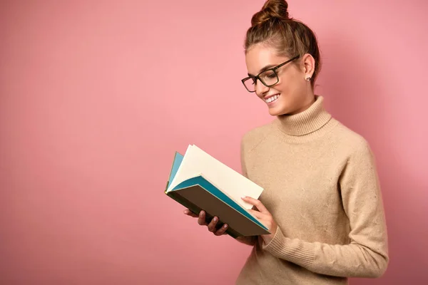 Egy lány, tiszta bőrrel és magas szemüveggel, félig megfordult állapotban áll, és mosolyogva néz a nyitott könyvre. — Stock Fotó