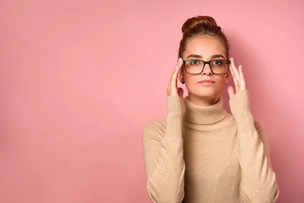Uma menina com pele limpa e um pão alto fica no fundo rosa e olha para a moldura, ajustando os óculos com os dedos . — Fotografia de Stock