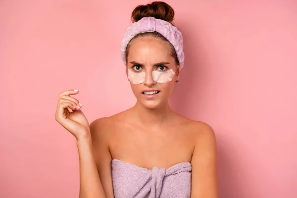 Seorang gadis dengan kulit bersih berdiri di atas latar belakang merah muda dengan handuk dan dengan tambalan di bawah matanya terlihat tidak menyenangkan di kamera . — Stok Foto