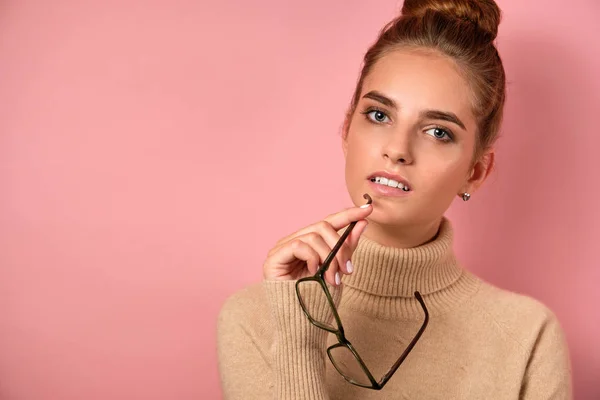 En flicka med ren hud och en hög bulle står på rosa bakgrund och ser eftertänksamt in i ramen, hålla glasögon vid örat. — Stockfoto