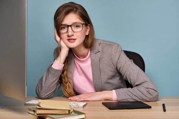 A girl in a pink sweatshirt and a gray jacket with a braid and glasses sits at a computer and leaning on hand looks into the frame — Stock Photo, Image