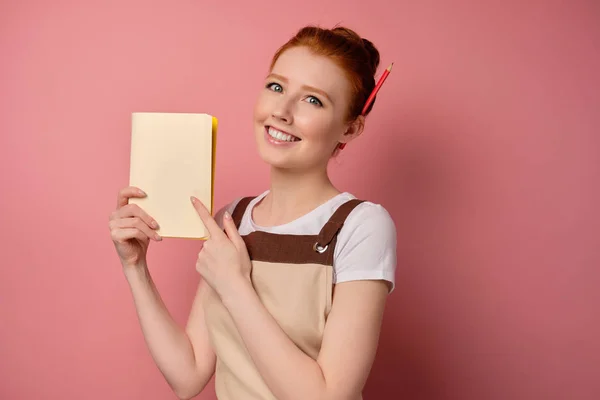 En söt rödhårig flicka med en hög bulle i ett beige förkläde som pekar på den bärbara sidan med finger och leende — Stockfoto