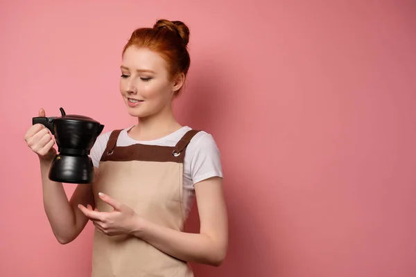 En söt rödhårig flicka med en hög bulle i ett beige förkläde står på en rosa bakgrund och tittar på kaffekannan i handen — Stockfoto