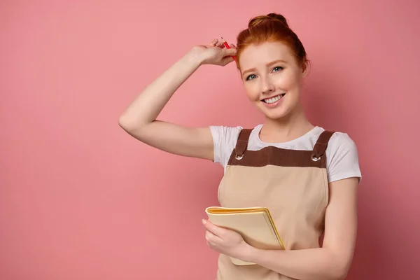 En söt rödhårig flicka med en hög bulle i ett beige förkläde ler, håller en anteckningsbok för sig själv och sätter en penna bakom örat — Stockfoto