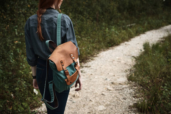 Рыжая девушка в синей рубашке с рюкзаком на плече идет по тропинке, рамка сзади, сосредоточьтесь на рюкзаке
.