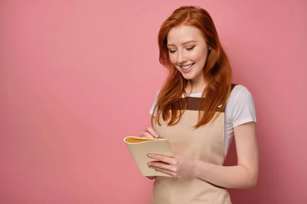 Den rödhåriga tjejen i ett beige förkläde och en T-shirt står på en rosa bakgrund leende och gör en anteckning i en anteckningsbok — Stockfoto