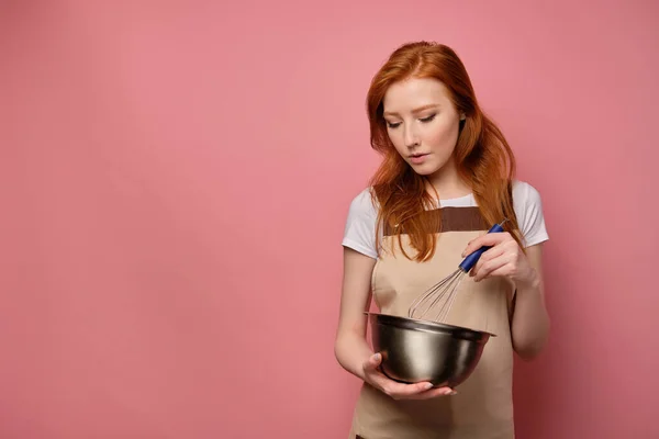 En rödhårig flicka i ett beige förkläde och en T-shirt står på en rosa bakgrund, piskar med en visp i en skål, bugar huvudet — Stockfoto