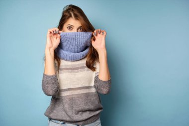 Süveter giymiş esmer bir kadın mavi bir arka planda duruyor ve yüzüne bir eşarp takıp çerçevenin içine bakıyor..