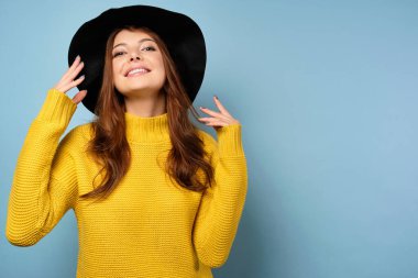 Sarı kazaklı ve şapkalı güzel bir esmer mavi arka planda duruyor, geniş bir gülümseme ve saçını düzleştiriyor.