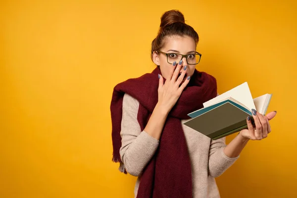 Egy barna nő, magas zsemlével, pulóverben és sálban áll, nyitott könyvet tart, és a kamerába néz, és kézzel takarja be az arcát. — Stock Fotó
