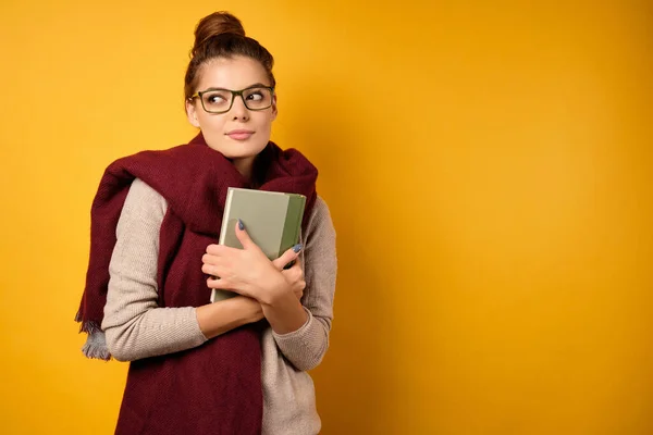 Egy gyönyörű barna nő, magas zsemlével és szemüveggel, pulóverben és sálban áll, a mellkasához nyom egy könyvet, oldalra néz. — Stock Fotó