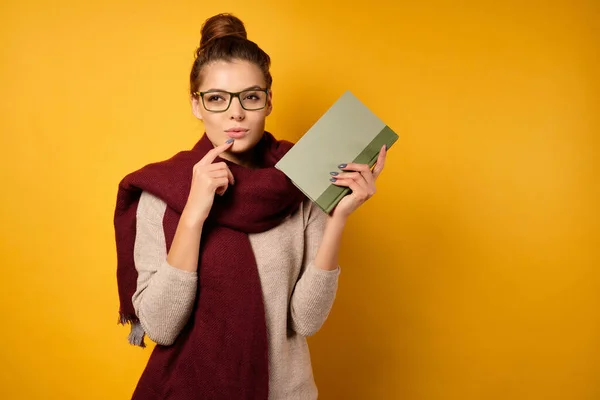 En brunett med en hög bulle och glasögon står i en tröja och halsduk, håller en bok i handen och ser sidledes med en skelning — Stockfoto