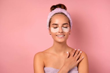 Temiz tenli bir kız pembe bir arka planda duruyor, gözleri kapalı ve gözlerinin altı yamalı..