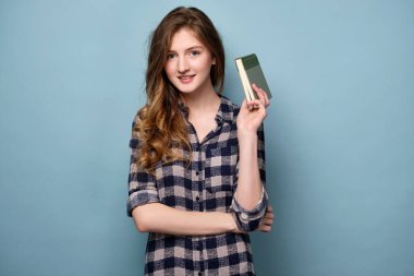 Ekose gömlekli bir kız mavi arka planda duruyor ve elinde bir kitap tutarken gülümsüyor.