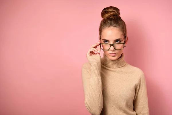 Uma menina com pele limpa e um pão alto fica no fundo rosa e parece descontente ao lado sobre os óculos . — Fotografia de Stock