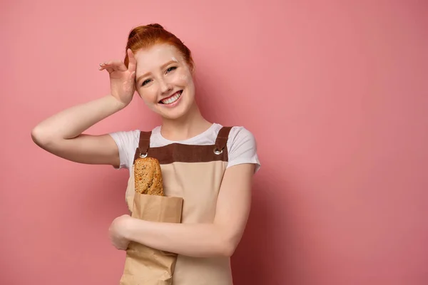 En söt rödhårig flicka med en hög bulle i ett beige förkläde, skrattar, tar bort slick mjöl och kramar en påse med en limpa — Stockfoto