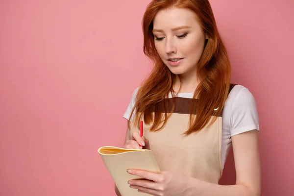 Rödhårig flicka i ett beige förkläde och en T-shirt står på en rosa bakgrund och gör en anteckning i en anteckningsbok. — Stockfoto