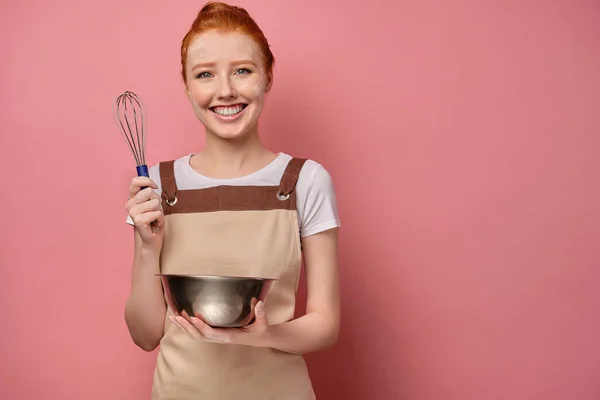 En söt rödhårig flicka med en hög bulle i ett beige förkläde och ett ansikte i mjöl ler brett, håller en visp och en skål i händerna — Stockfoto