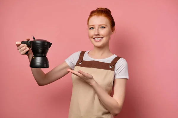 En söt rödhårig flicka med en hög bulle i ett förkläde ler in i ramen, visar kaffekannan i handen — Stockfoto