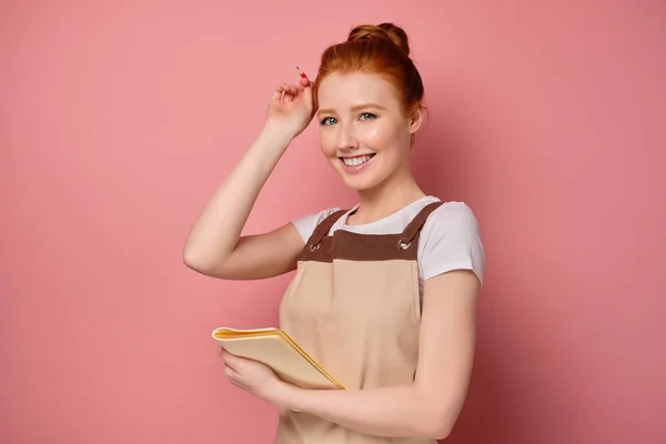 En söt rödhårig flicka med en hög bulle i ett beige förkläde, håller i en anteckningsbok och sätter en penna bort från örat. — Stockfoto