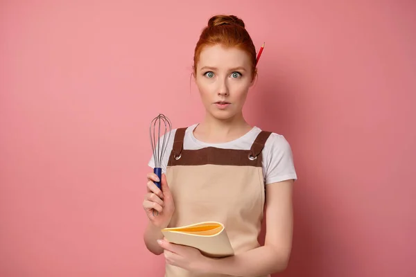 En söt rödhårig flicka med en hög bulle i ett beige förkläde ser förvånad ut i ramen, håller en anteckningsbok och en visp i händerna — Stockfoto