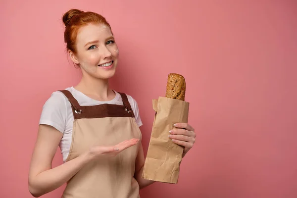 En söt rödhårig flicka med en hög bulle i ett beige förkläde, med ansiktet i mjöl och leende, pekar på ett paket med en limpa — Stockfoto