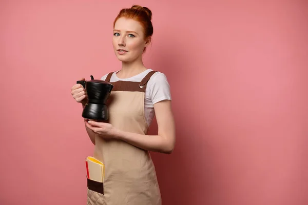 En söt rödhårig flicka med en hög bulle i ett beige förkläde står på en rosa bakgrund, med en kaffekanna i händerna — Stockfoto