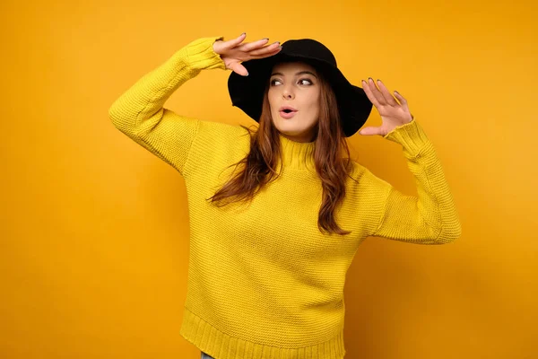 Na žlutém pozadí stojí bruneta ve žlutém svetru a černý klobouk se založenýma rukama, hledí do strany — Stock fotografie