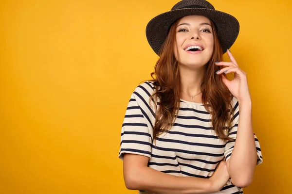 Eine schöne Brünette in gestreiftem T-Shirt und Korbflechter steht auf gelbem Grund, lacht, hebt das Kinn. — Stockfoto