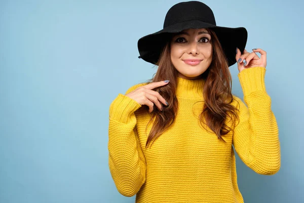 Uma morena em um suéter amarelo e chapéu fica em um fundo azul de surpresa com olhos redondos e segurando a borda do chapéu. — Fotografia de Stock