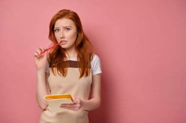 Önlüklü kızıl saçlı kız elinde bir defterle elinde kalemle çerçeveye endişeyle bakıyor.